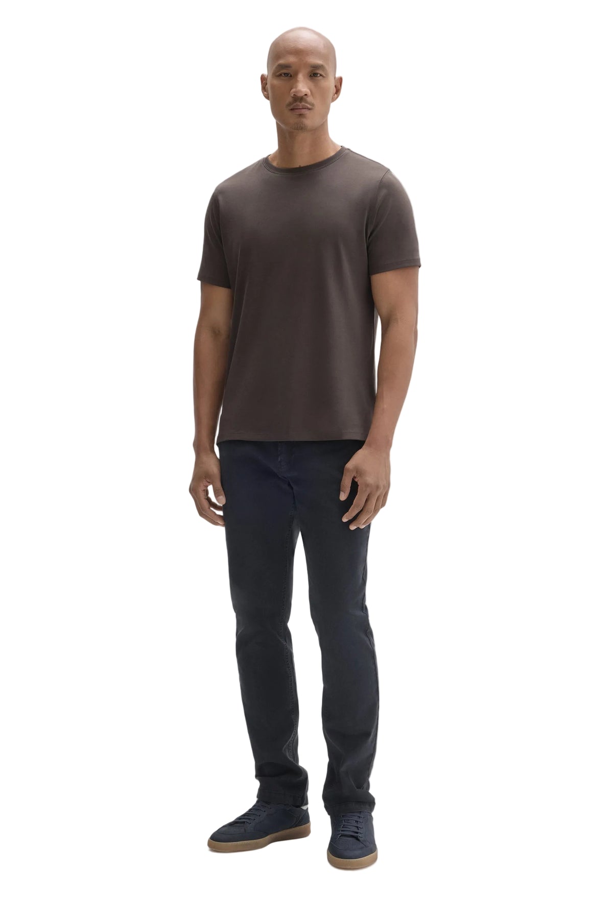 A man modeling a dark brown short-sleeve crewneck t-shirt, dark navy blue jeans, and navy blue slip-on shoes against a solid light gray background.
