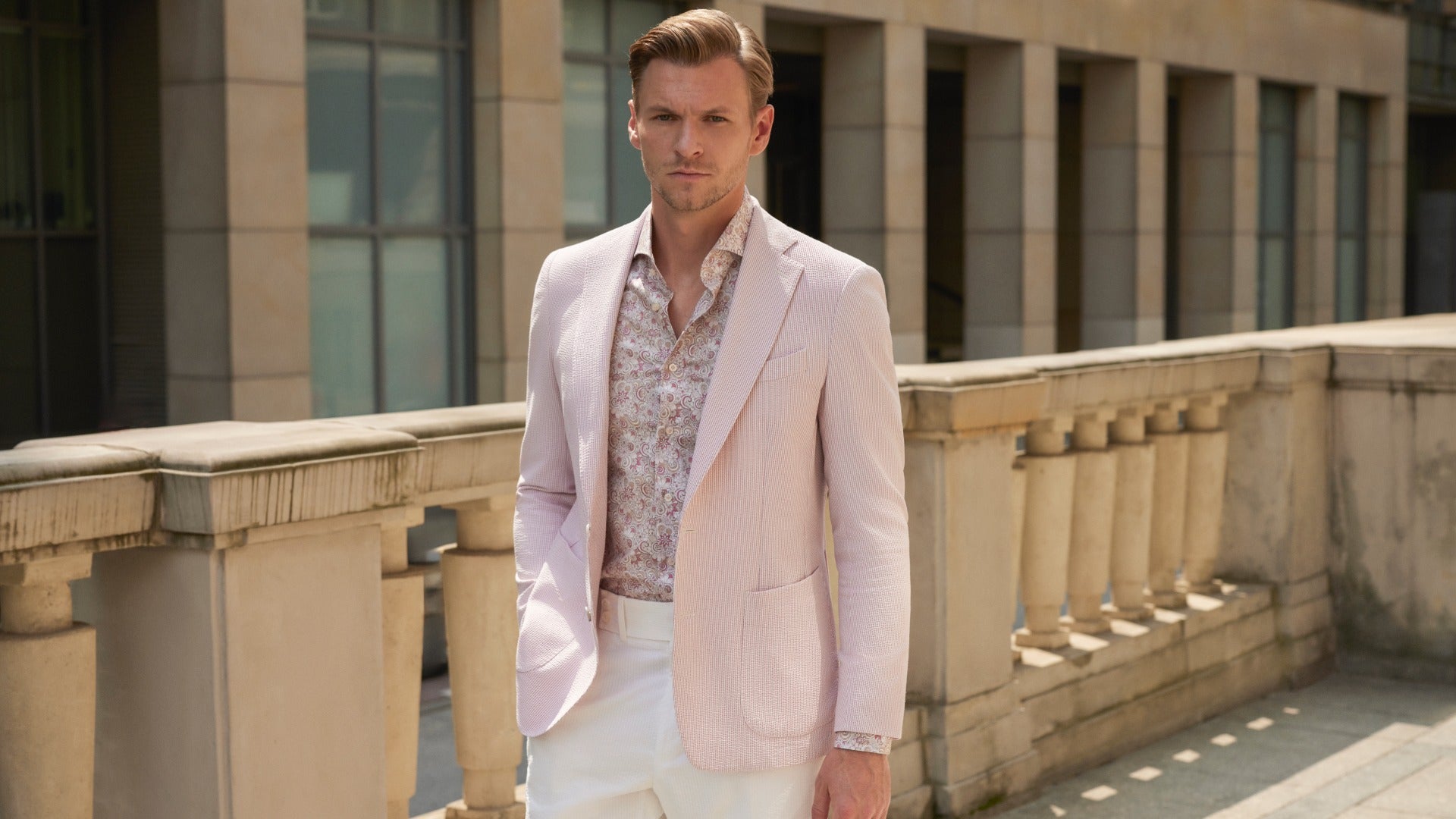 Man in light pink blazer and patterned shirt standing on a stone balcony.