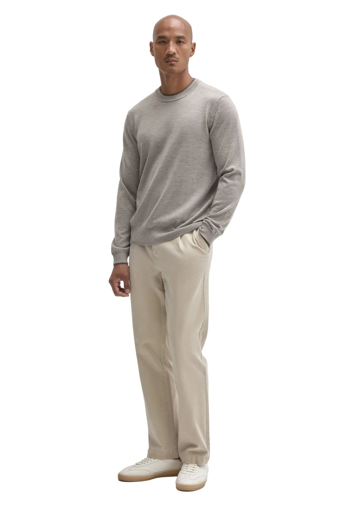 A man modeling a light gray long-sleeve crewneck sweater, beige trousers, and off-white sneakers against a solid light gray background.